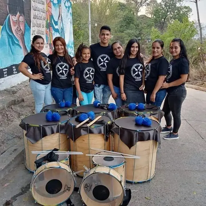 Jóvenes integrantes de una batucada posan juntos frente a un mural comunitario